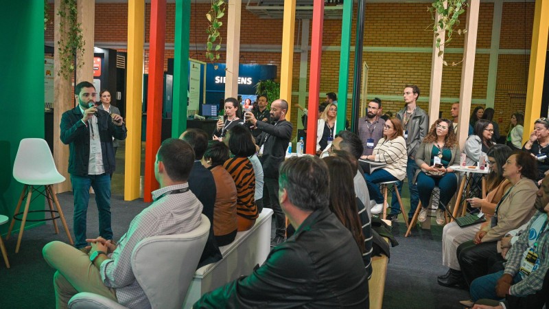 Foto que enquadra a plateia de lado, com Gabriel à frente falando ao microfone em pé. Há muitas pessoas sentadas e em pé no evento.